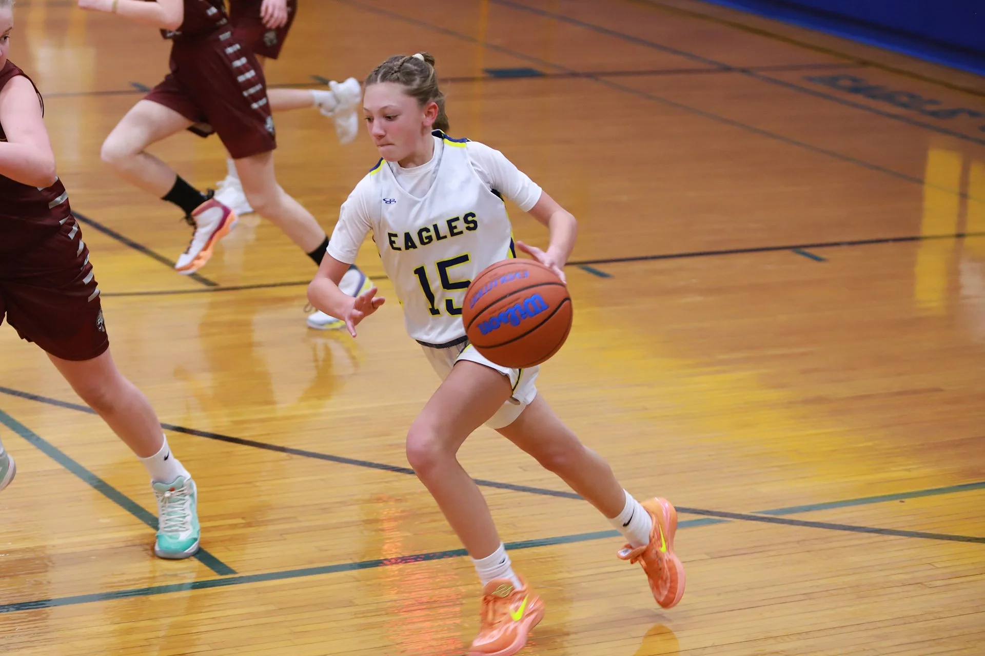 A young girl in a white basketball uniform with the number 15 and the word 'Eagles' on her jersey dribbles a basketball up the court.