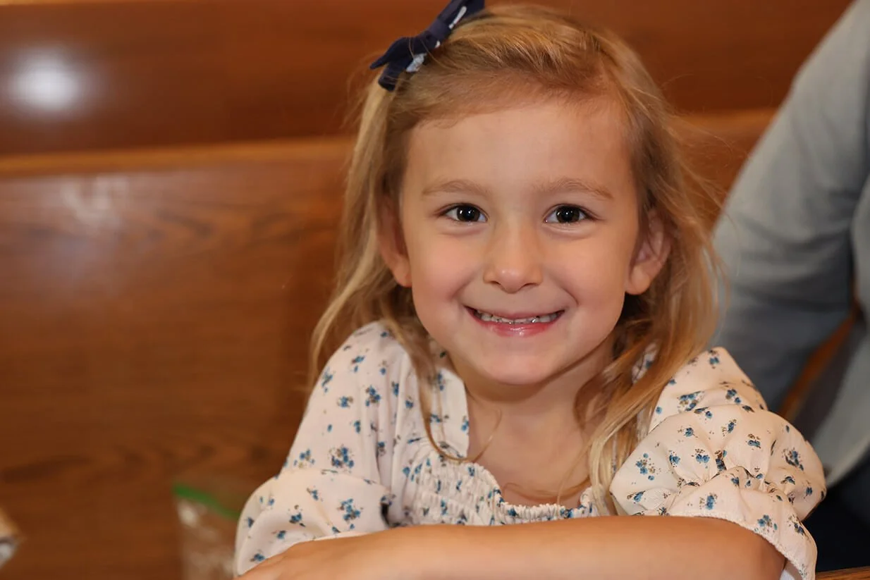 A smiling young girl with red hair, wearing a floral blouse and a navy blue bow, sitting at a wooden table in an indoor setting.