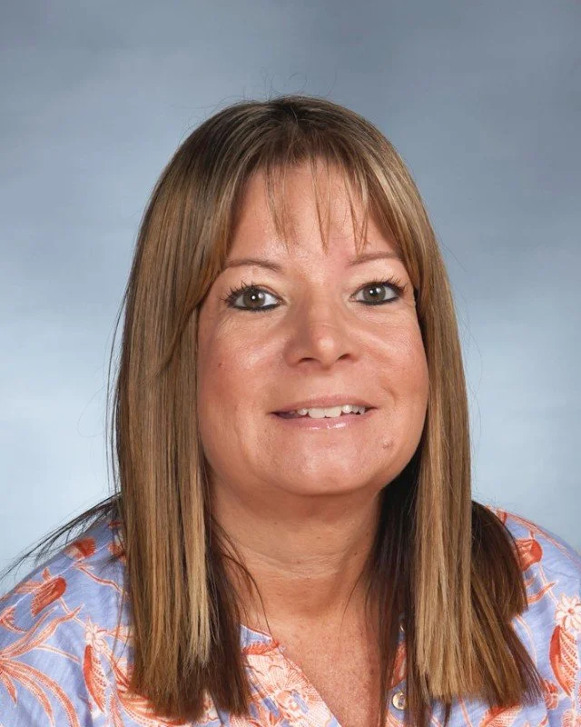 Portrait of a woman with light brown hair and brown eyes, wearing a blue shirt with orange and white floral pattern, smiling at the camera.