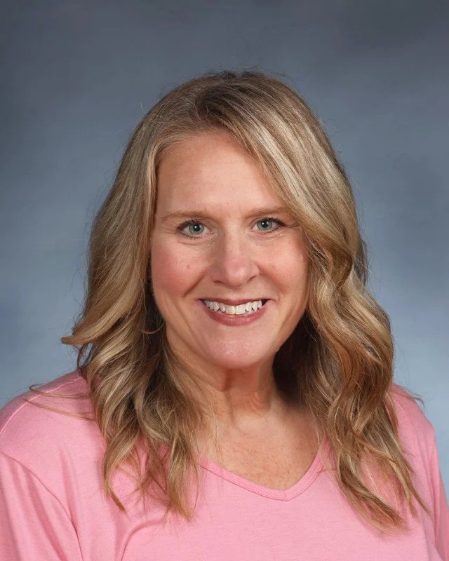 Smiling woman with wavy blonde hair wearing a pink top, posed in front of a neutral background.