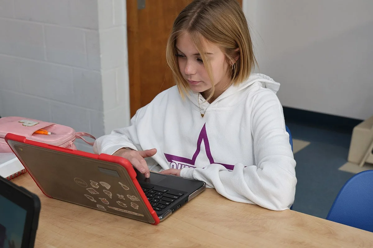 A young girl with blonde hair wearing a white hoodie with a purple star design, sitting at a desk and using a laptop with stickers on it.