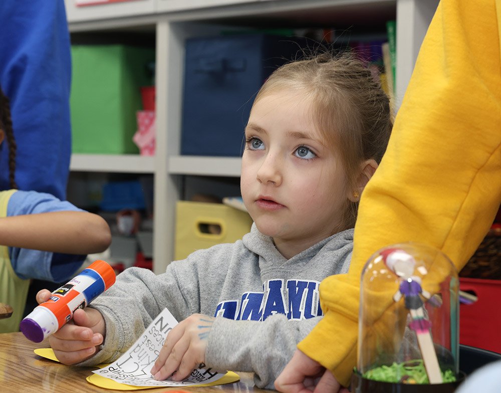 A young girl with blue eyes and light brown hair tied back, wearing a gray hoodie, sitting at a table in a classroom or activity setting. She is holding a glue stick and looking attentively at something off-camera. There are colorful storage bins in the background and a small craft project on the table.