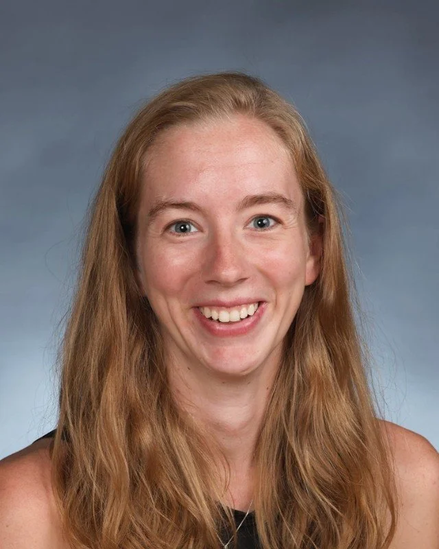 A smiling woman with long red hair, blue eyes, and fair skin against a neutral background.