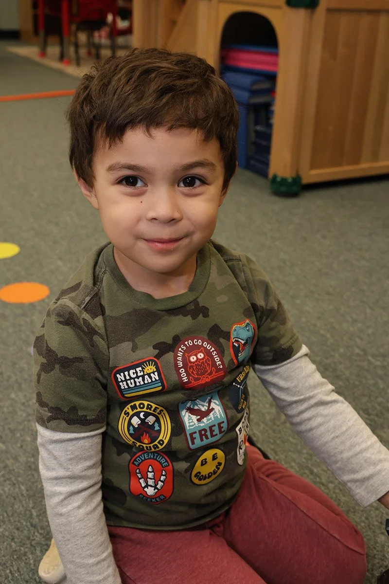 A young boy with short brown hair and brown eyes, smiling at the camera. He is wearing a camouflage shirt with various colorful patches and maroon pants, sitting on a playroom or classroom carpet.