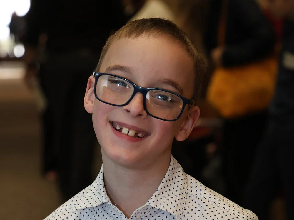 A young boy with glasses smiling, showing missing teeth, wearing a white button-up shirt with small black polka dots.