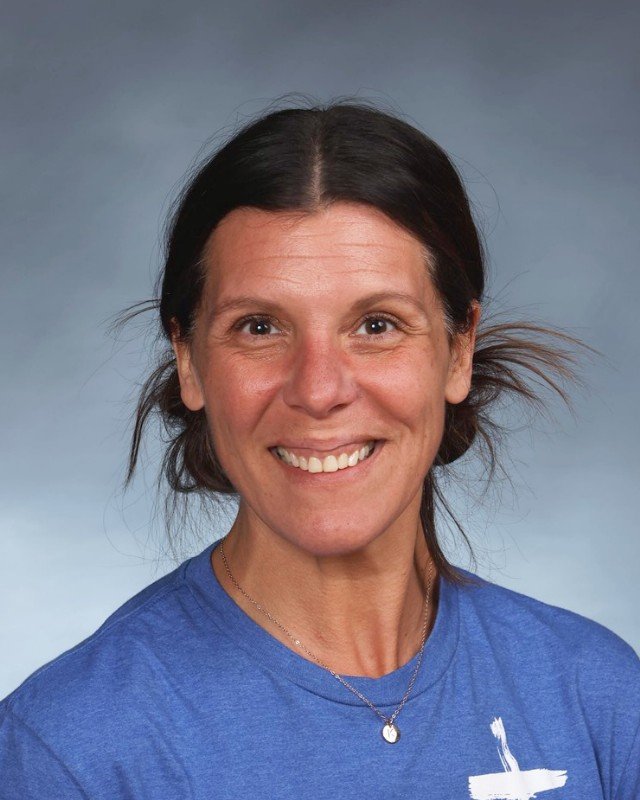 A woman with dark hair smiling, wearing a blue shirt and a necklace against a gray background.