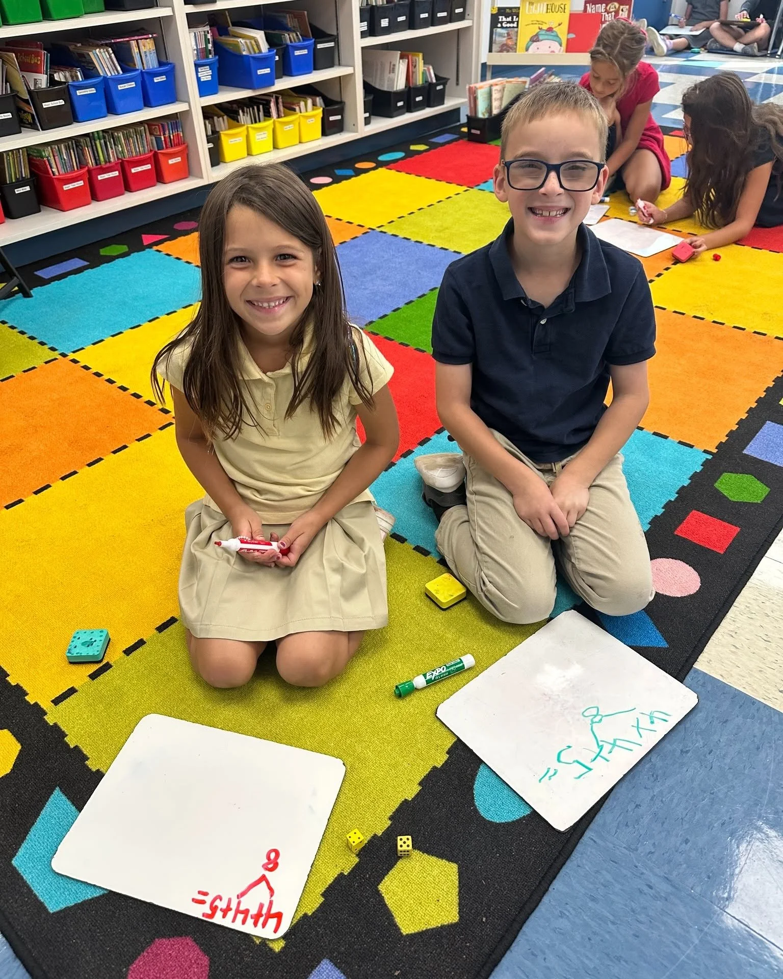 Two children, a girl and a boy, sitting on a colorful classroom rug, smiling at the camera.