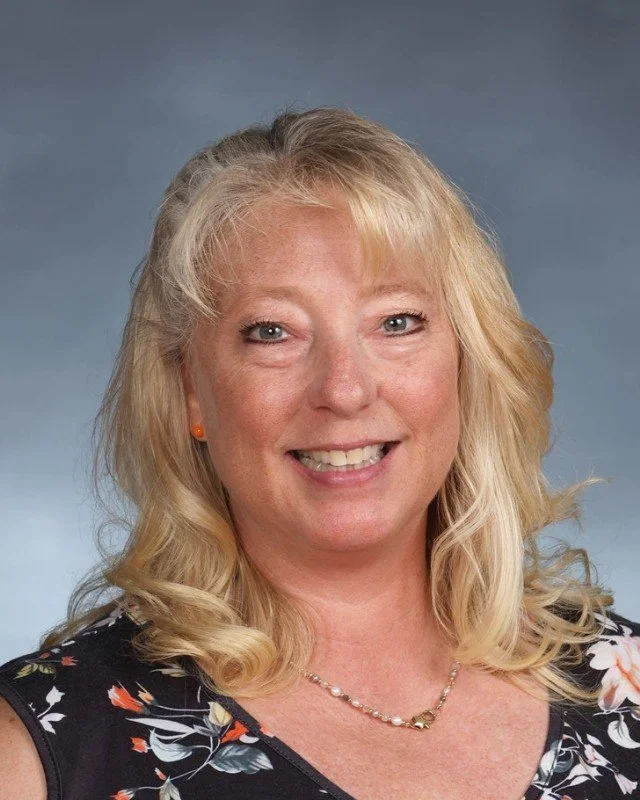 A woman with blonde hair and blue eyes smiling at the camera, wearing an orange earring, a pearl necklace, and a black floral dress.