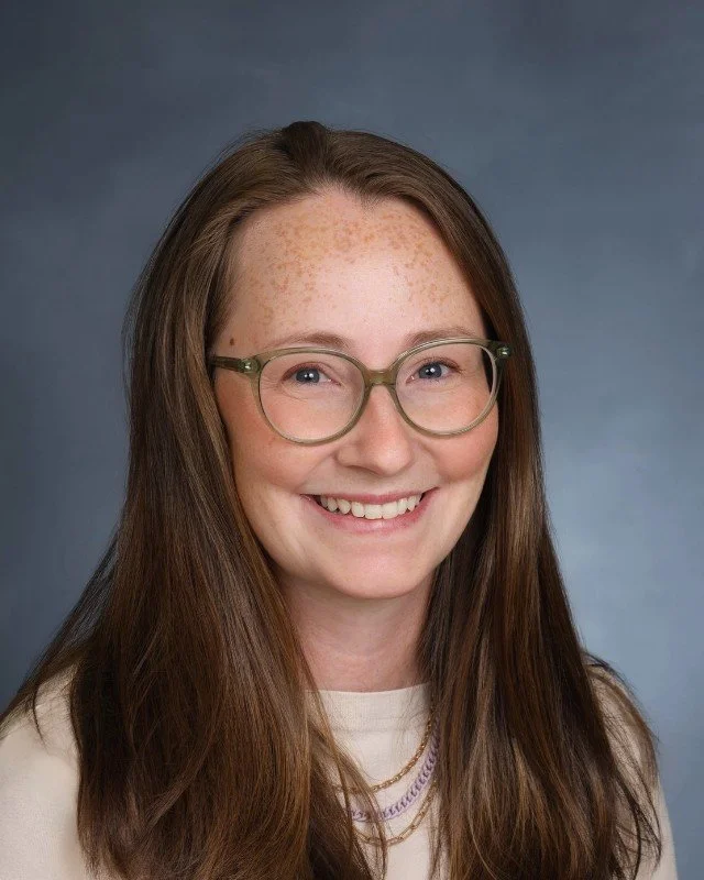A young woman with long brown hair, glasses, and freckles smiling against a blue background.