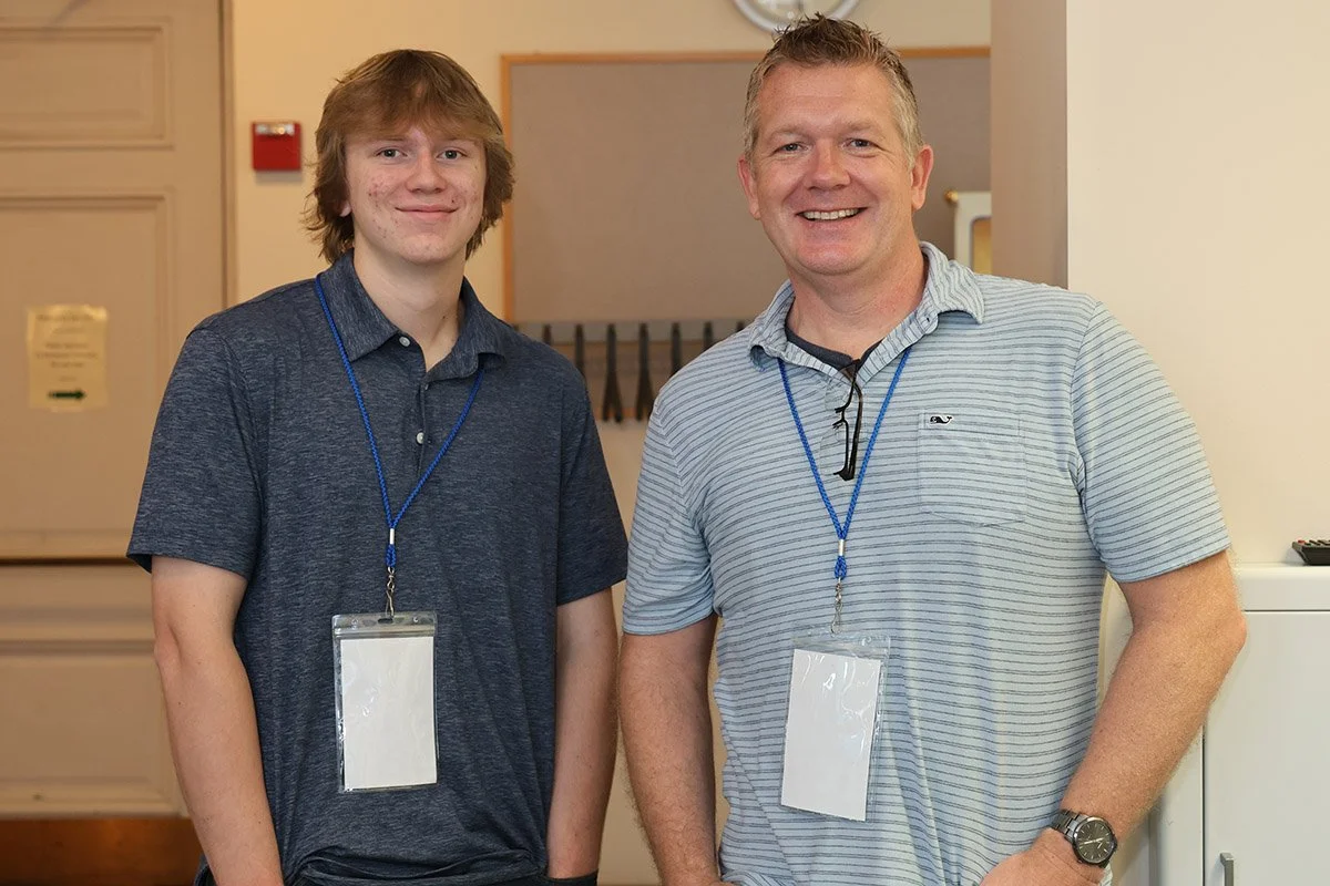 Two men standing indoors, both wearing blue lanyards with clear ID badges, smiling at the camera.