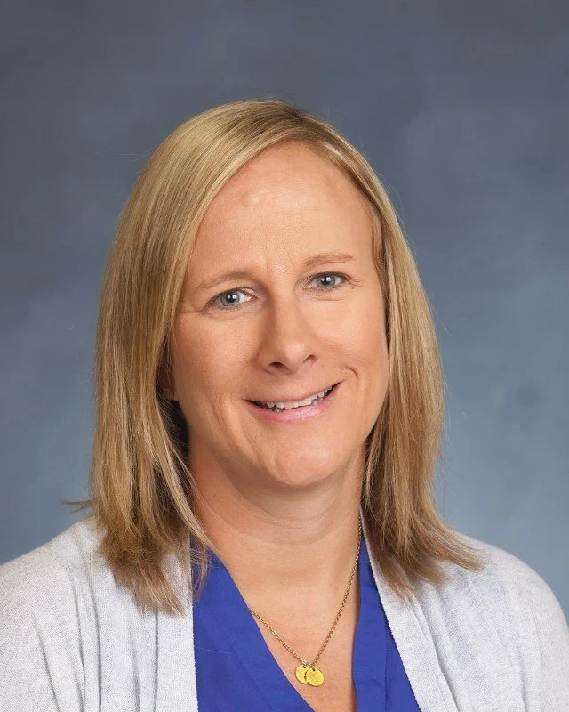 Portrait of a woman with shoulder-length blonde hair, blue eyes, wearing a blue top, gray cardigan, and a gold heart-shaped necklace, smiling against a plain blue background.