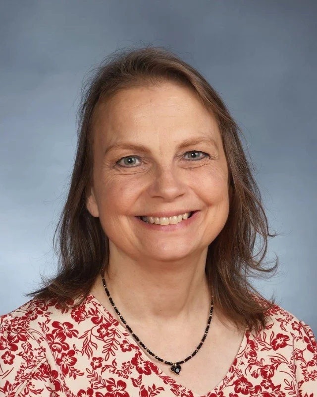 A woman with shoulder-length brown hair, blue eyes, and a friendly smile, wearing a red and white floral blouse and a black beaded necklace, against a plain blue background.