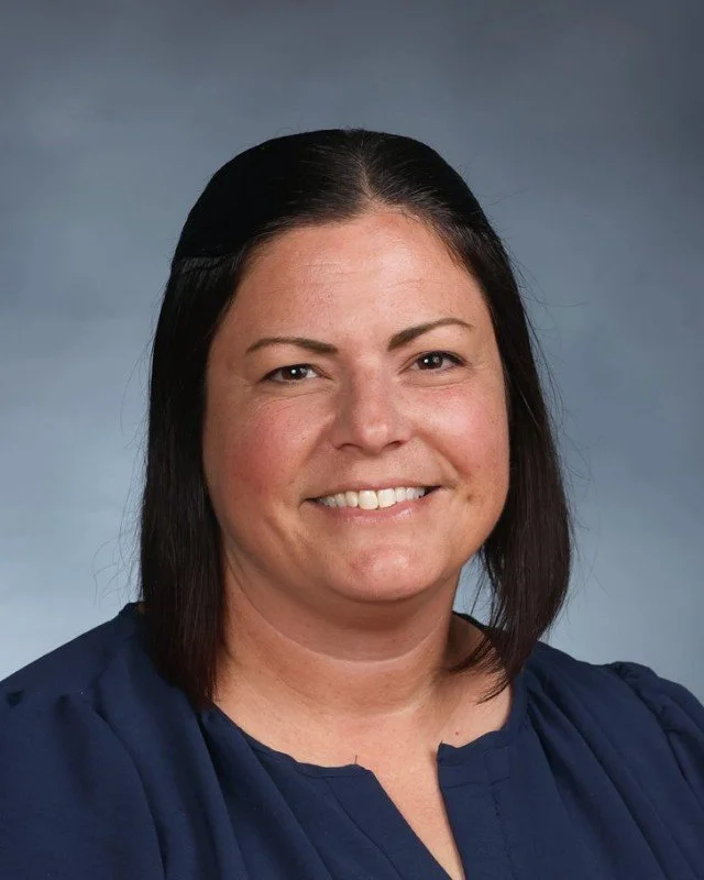 A woman with shoulder-length dark brown hair, wearing a navy blue blouse, smiling at the camera against a neutral gray background.