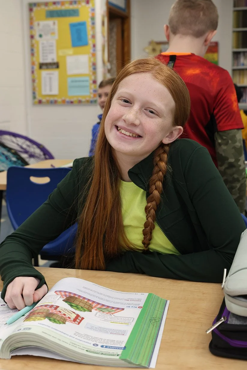 A smiling red-haired girl in a classroom, sitting at a desk with an open textbook in front of her, with other children and classroom decorations in the background.