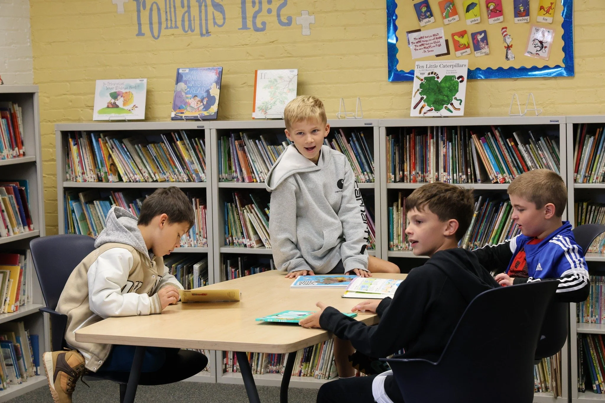 A group of children sitting and standing around a table in a library, with one boy standing and talking, and others reading or engaging with books.