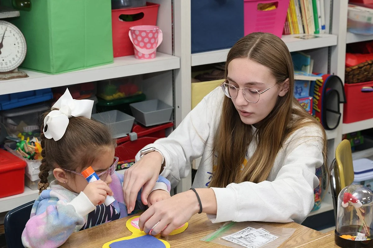 A young woman and a girl working together on a craft project at a table in a classroom. The girl has a large white bow in her hair and is holding a glue stick, while the woman is helping with the project.