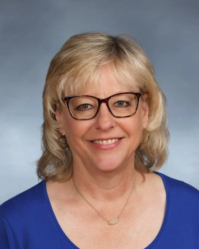 A woman with blonde hair, glasses, and a blue top smiling against a neutral background.