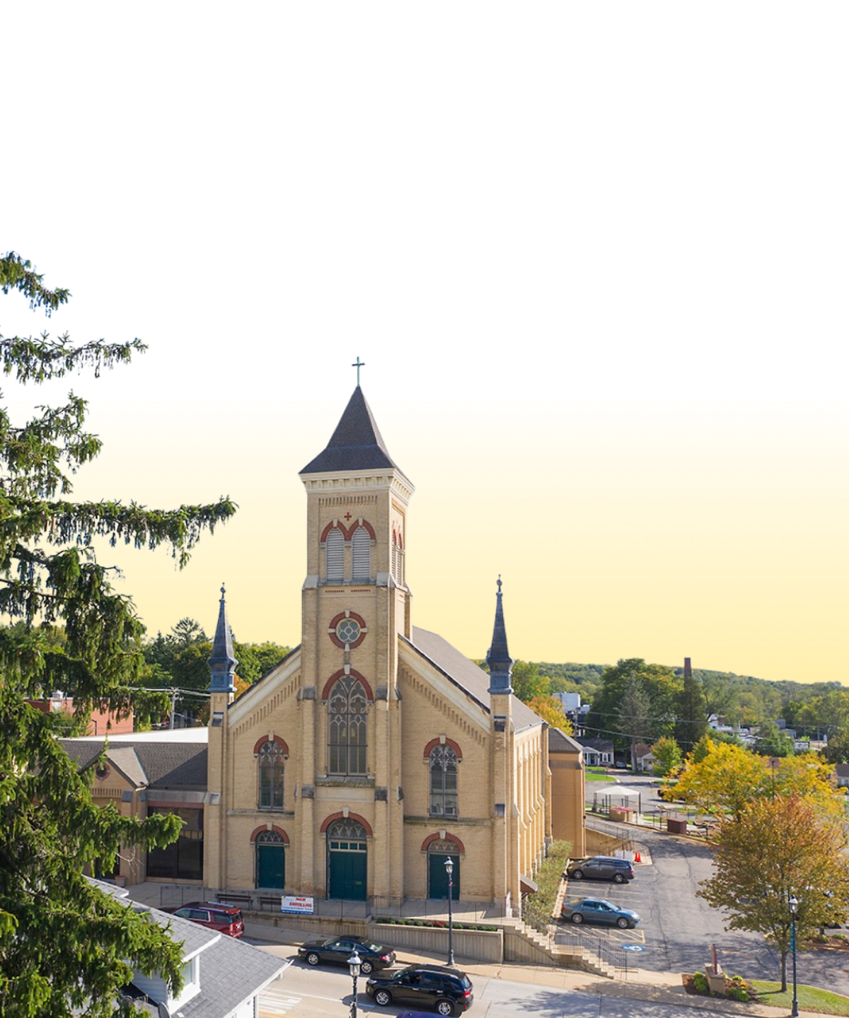 A historic church with a tall steeple and two smaller spires, surrounded by trees and parked cars, with a clear sky in the background.