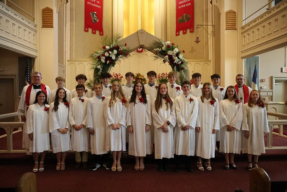 A group of young confirmands in white robes with red accents, standing on the steps of a church altar.