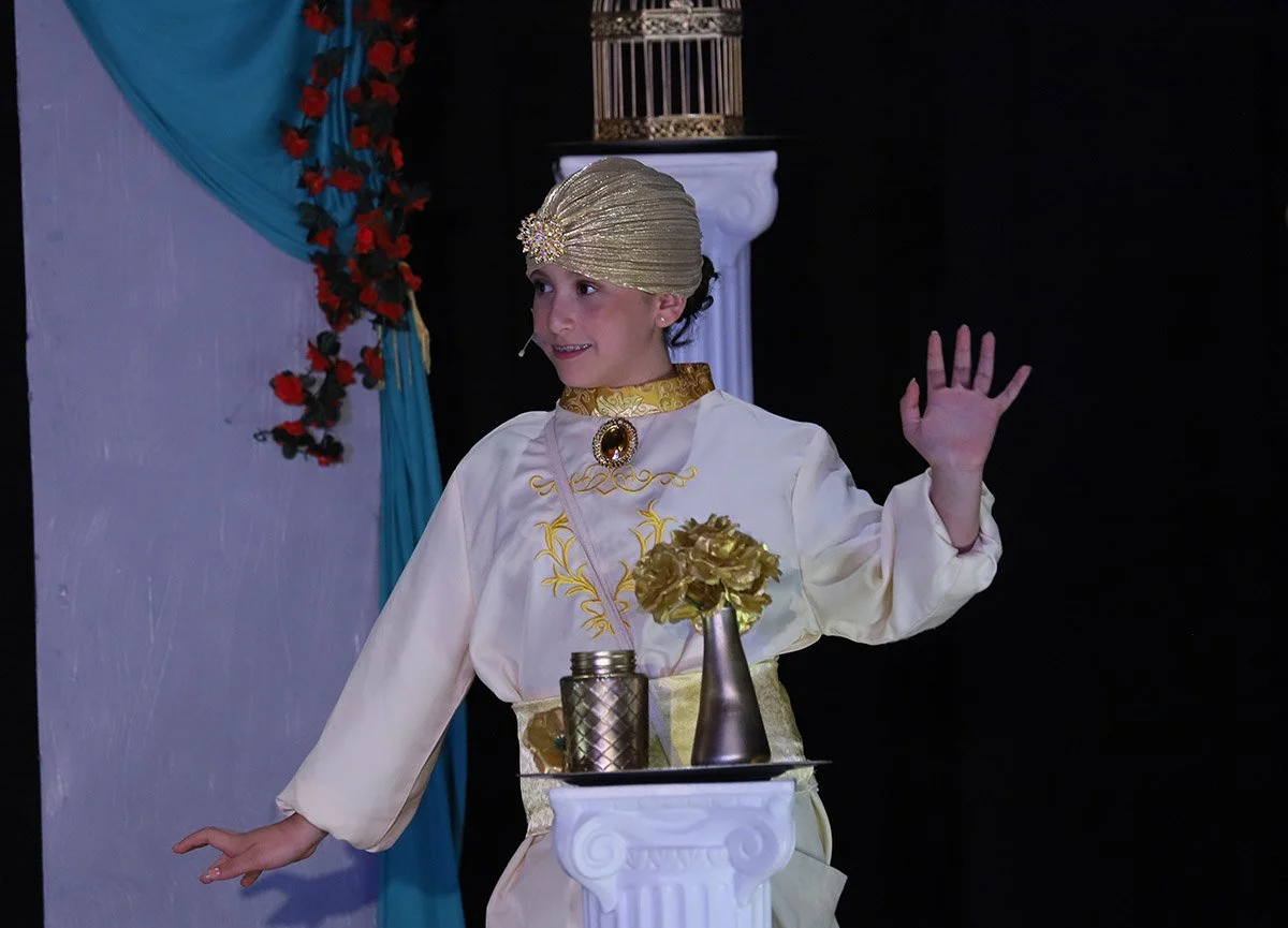 A young girl dressed in a costume with gold and white colors, wearing a gold turban with a decorative brooch, standing on a stage and waving her hand. The stage is decorated with a blue curtain, red flowers, and a white pedestal with gold accents.