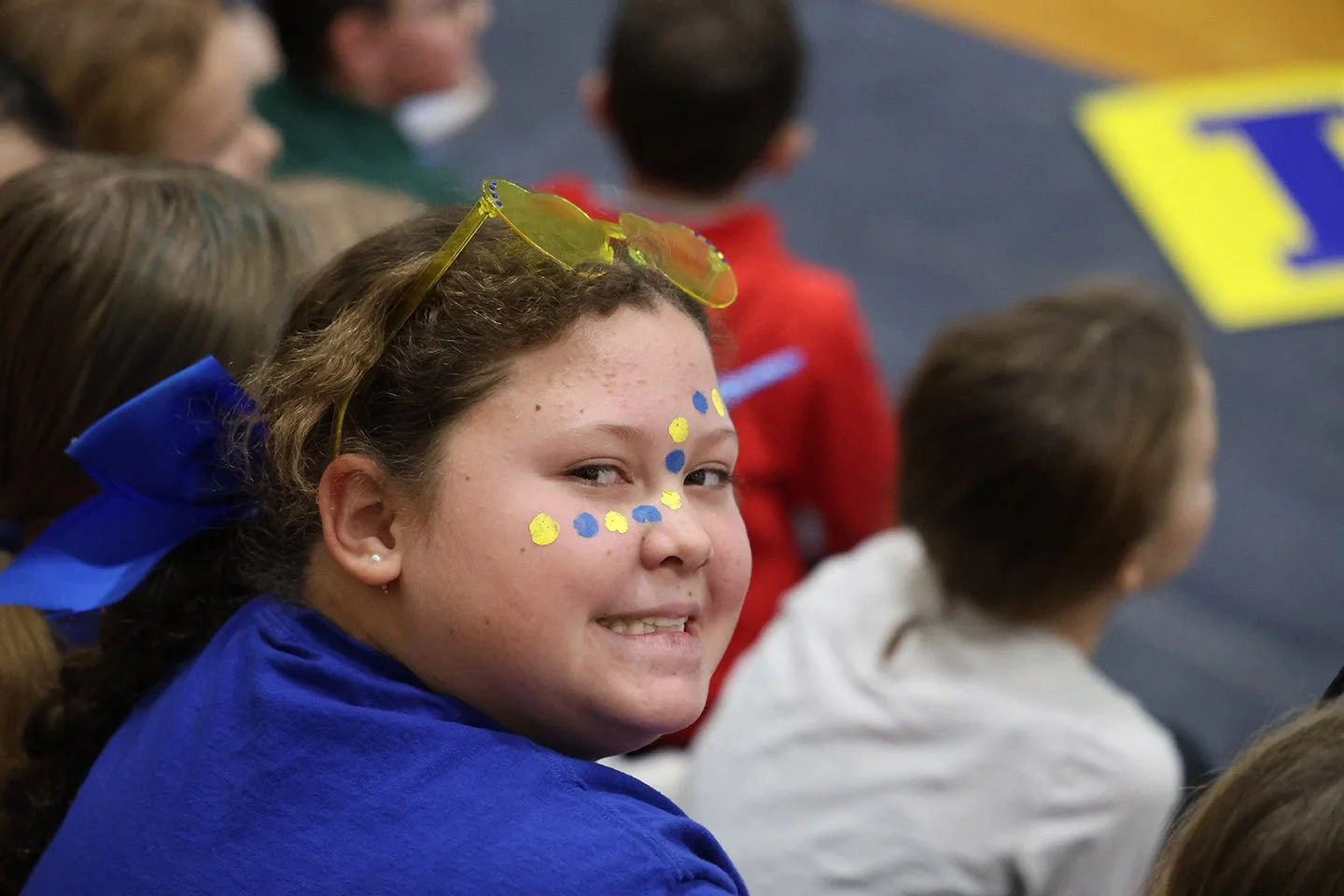 Smiling girl with face paint, a blue bow in her hair, and yellow-tinted glasses on her head, sitting among other children in a gym.