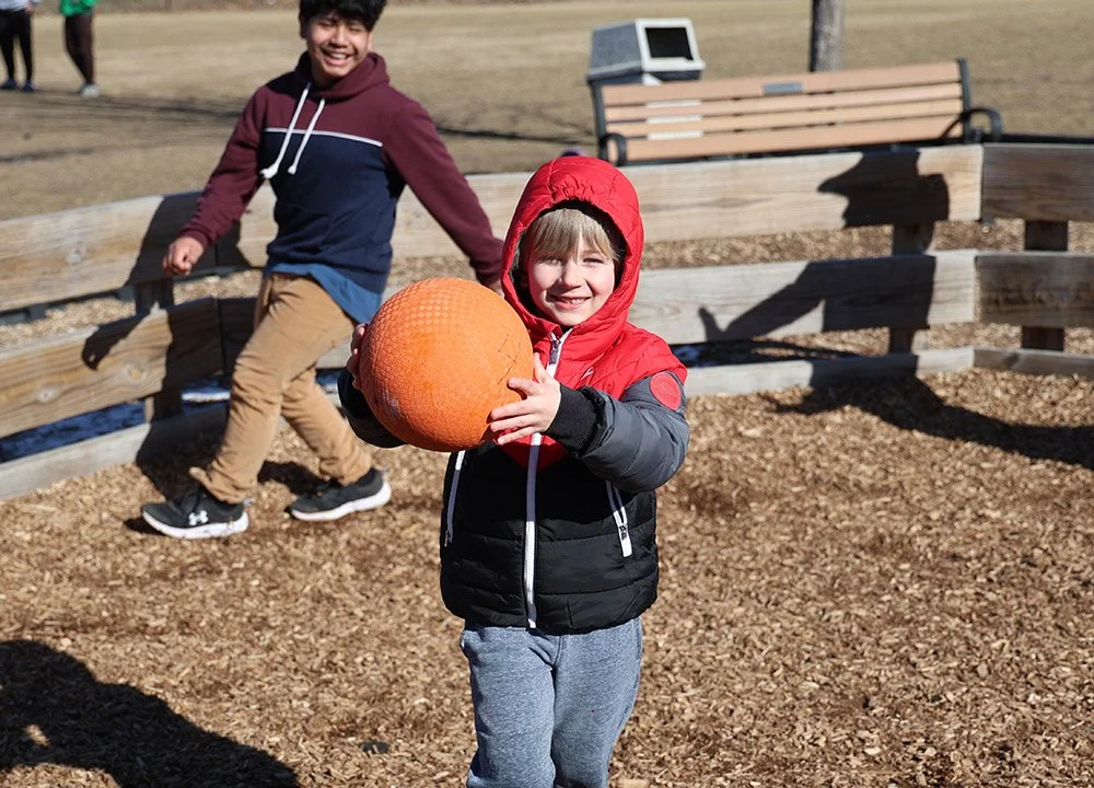Two kids playing outdoors, one child in a black and red jacket holding an orange basketball, the other in a maroon hoodie, with a wooden fence, a bench, and a dry field in the background.