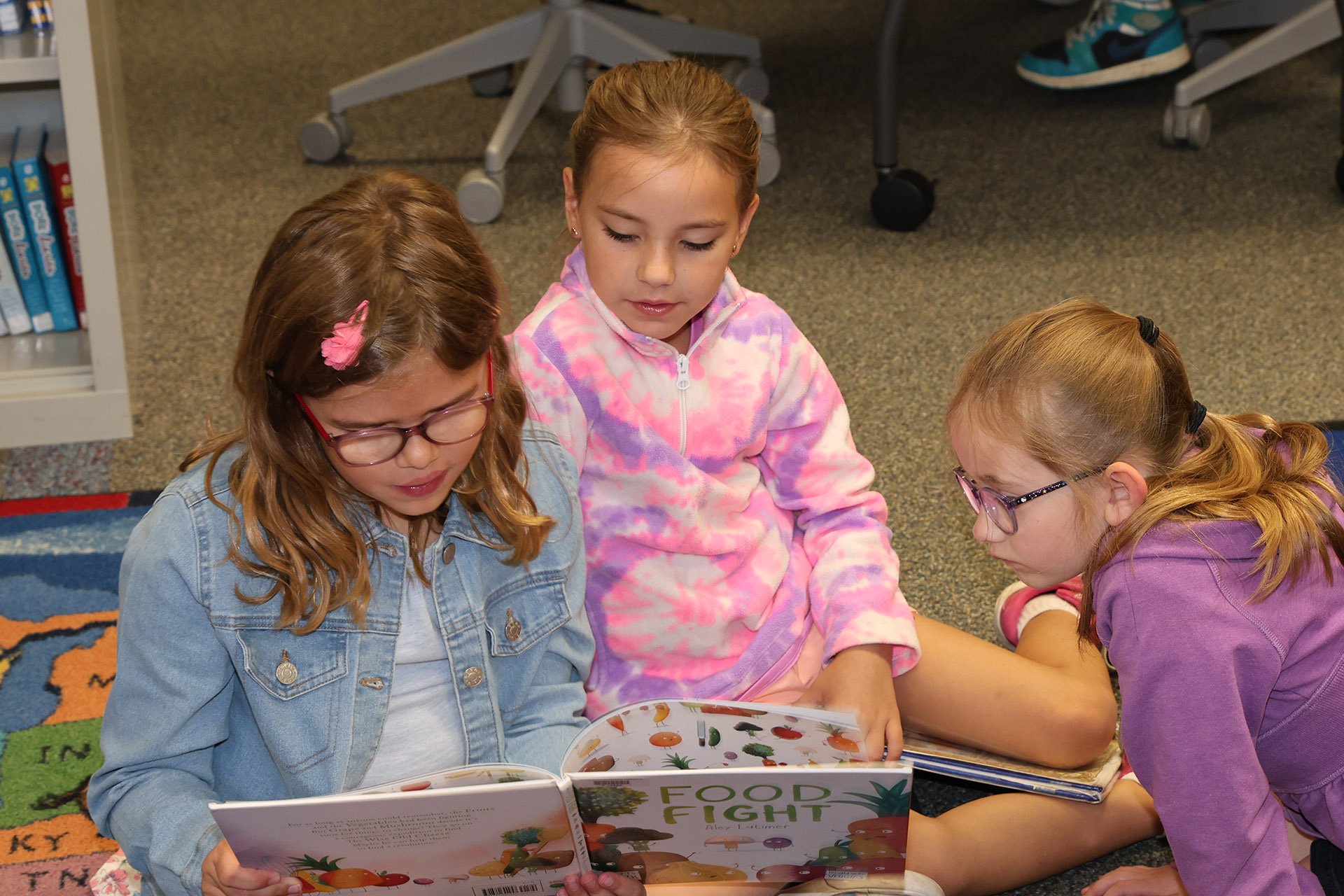 Three young girls sitting on the floor in a library, reading a colorful book titled 'Food Fight' together.