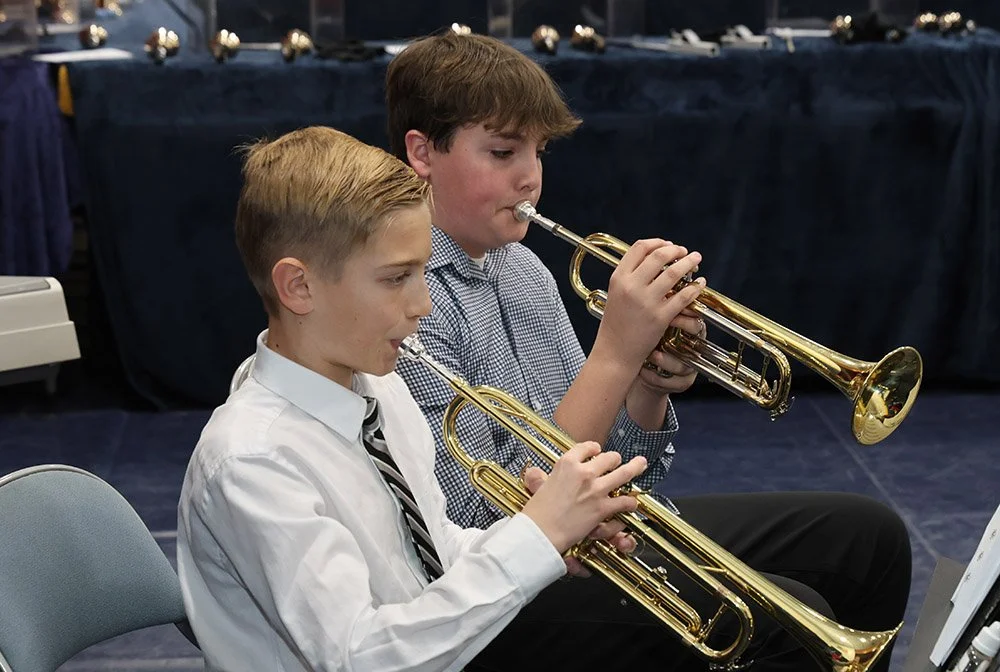 Two young boys in white shirts and ties playing trumpets in an indoor setting.