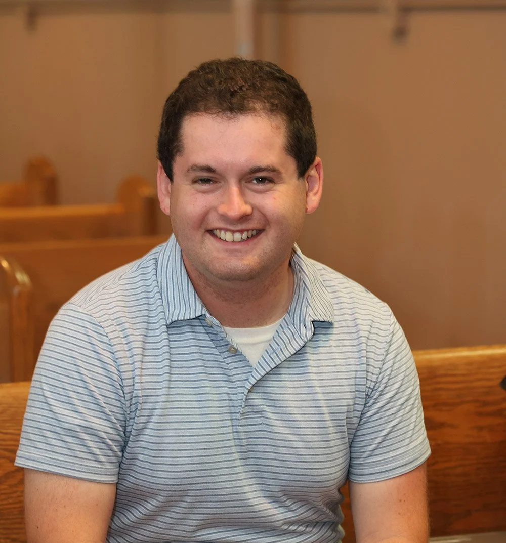 A smiling young man with short dark hair, wearing a light blue striped polo shirt, seated in a room with wooden chairs and a beige wall.