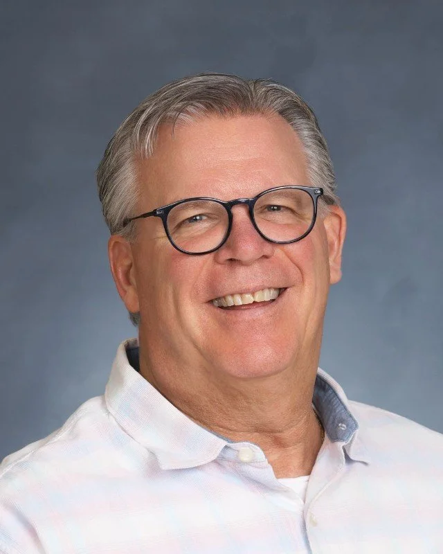 Portrait of a man with gray hair and glasses smiling, wearing a white collared shirt against a grey background.