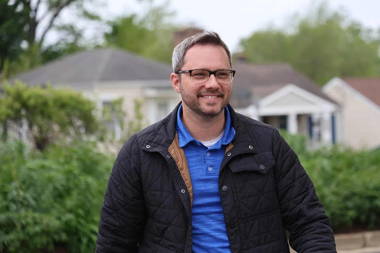 A man wearing glasses, a blue collared shirt, and a black quilted jacket standing outdoors with houses and greenery in the background.