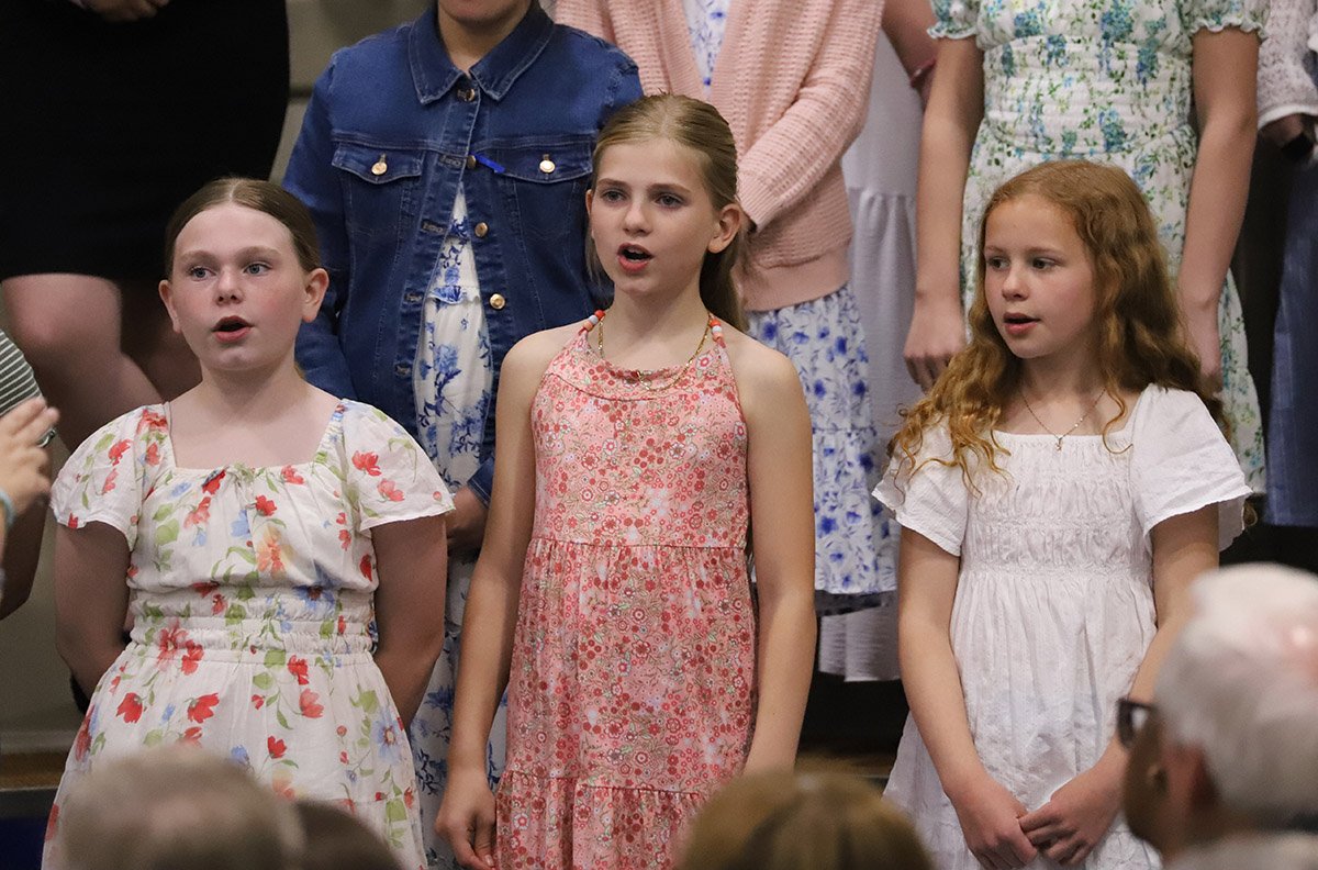 Three young girls are standing and singing at a children's choir performance.
