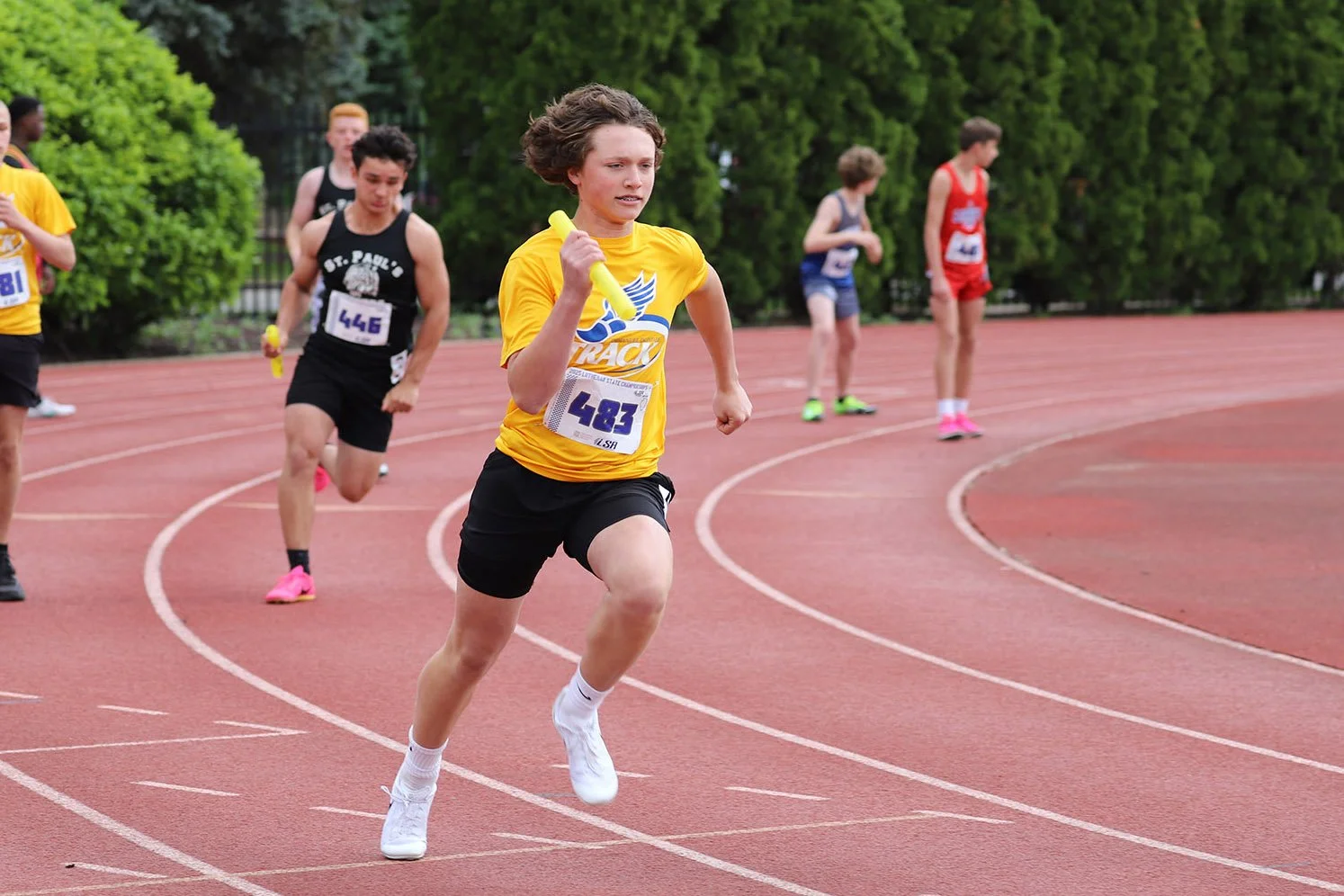 Young male athletes running on a red track during a race, with trees in the background.