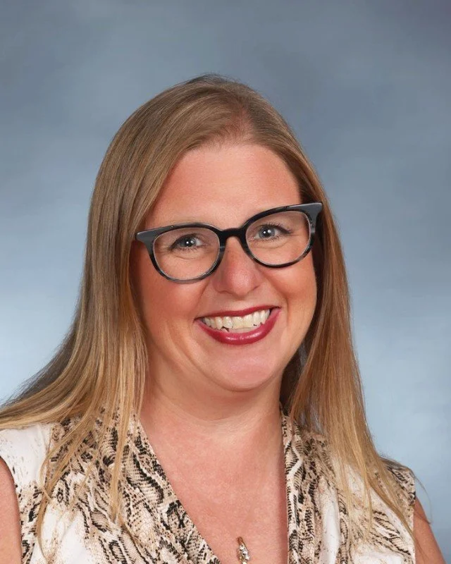A woman with long blonde hair, wearing glasses, smiling, and dressed in a sleeveless patterned top, posed against a neutral background.