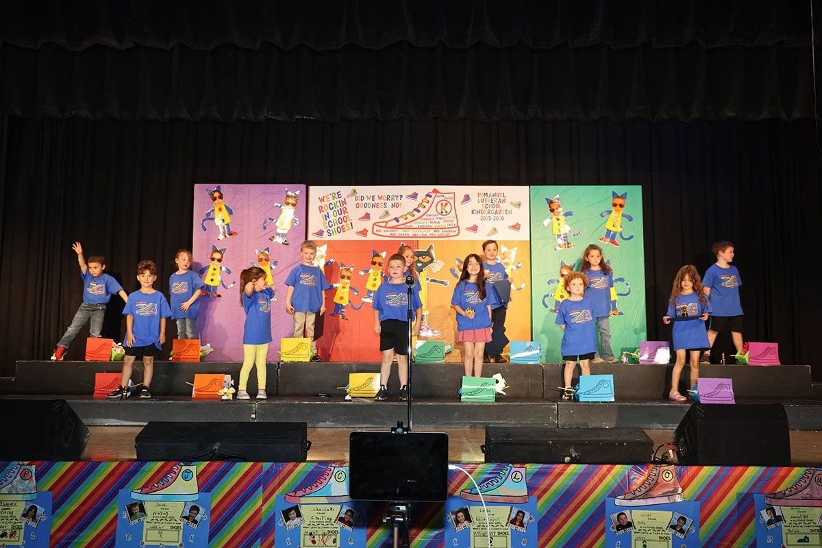 Children performing on stage at a school event, wearing matching blue T-shirts, with colorful decorations and banners in the background.