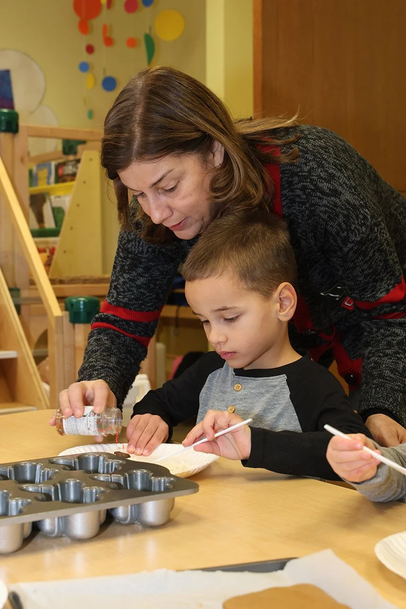 A woman assists a young boy in decorating cookies with sprinkles in a classroom or playroom setting.