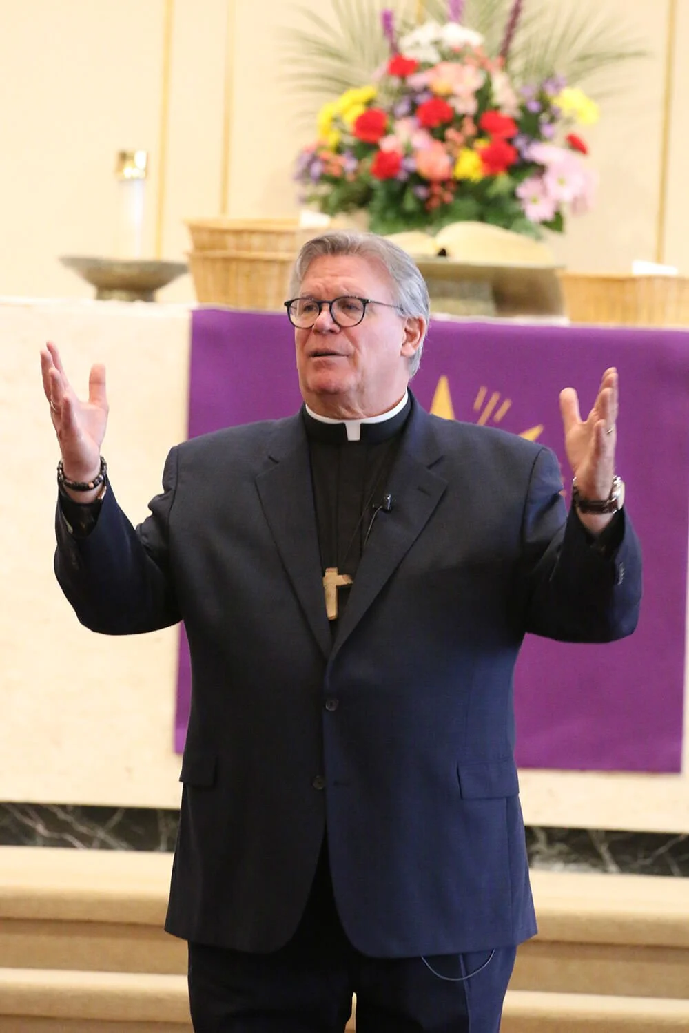 A man wearing a clerical shirt and cross necklace, speaking inside a church, with purple cloth and a floral arrangement in the background.