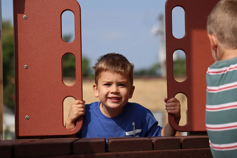 Two young boys are at a playground; one boy, wearing a blue shirt, is looking at the camera and holding onto the play structure, while the other boy, in a striped shirt, has his back to the camera.