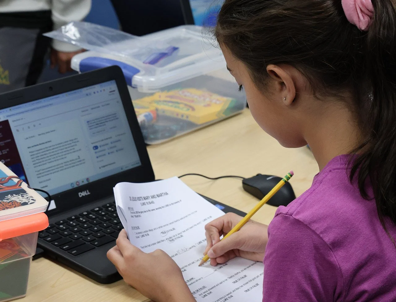 A young girl in a purple shirt sitting at a desk, writing on a paper with a yellow pencil, with an open laptop, mouse, and plastic storage containers on the desk.