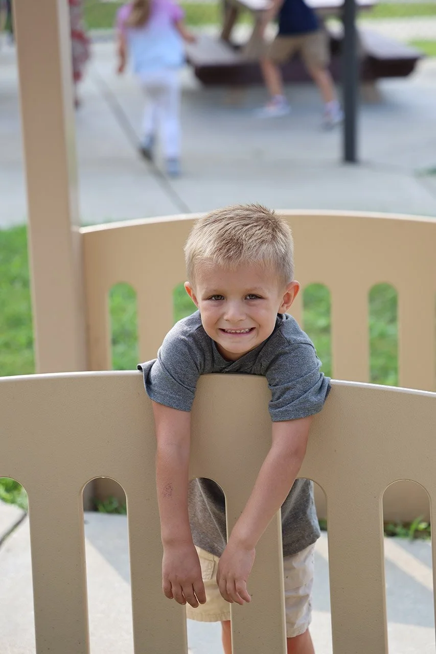 A young boy leaning on a beige gate at playground, smiling at camera, with children playing in the background.