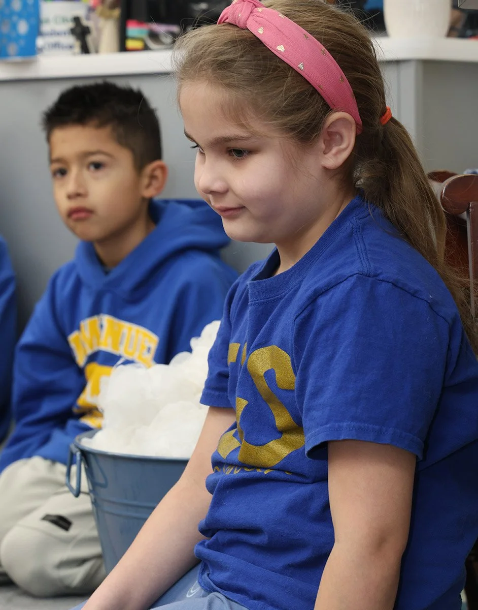 A young girl with a pink headband sitting in a classroom, with a boy in a blue hoodie.