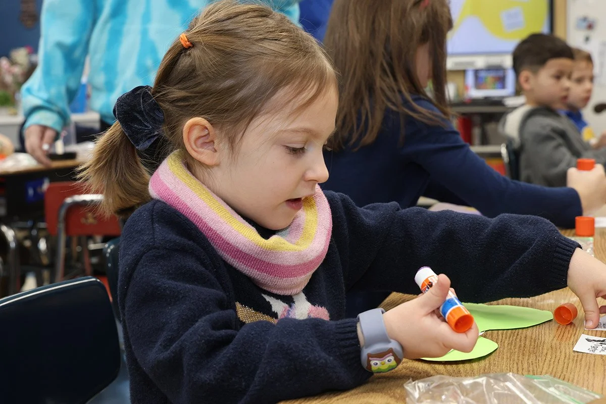 A young girl with brown hair in pigtails using glue stick on craft projects in a classroom.