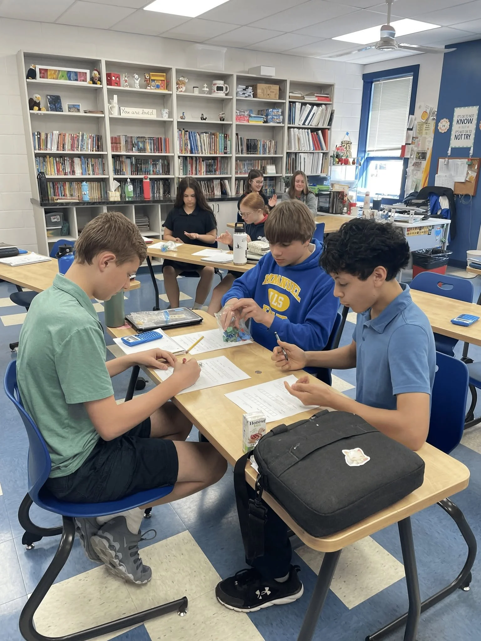 Students sitting at desks in a classroom, working on assignments, with a bookshelf filled with books and educational materials in the background.