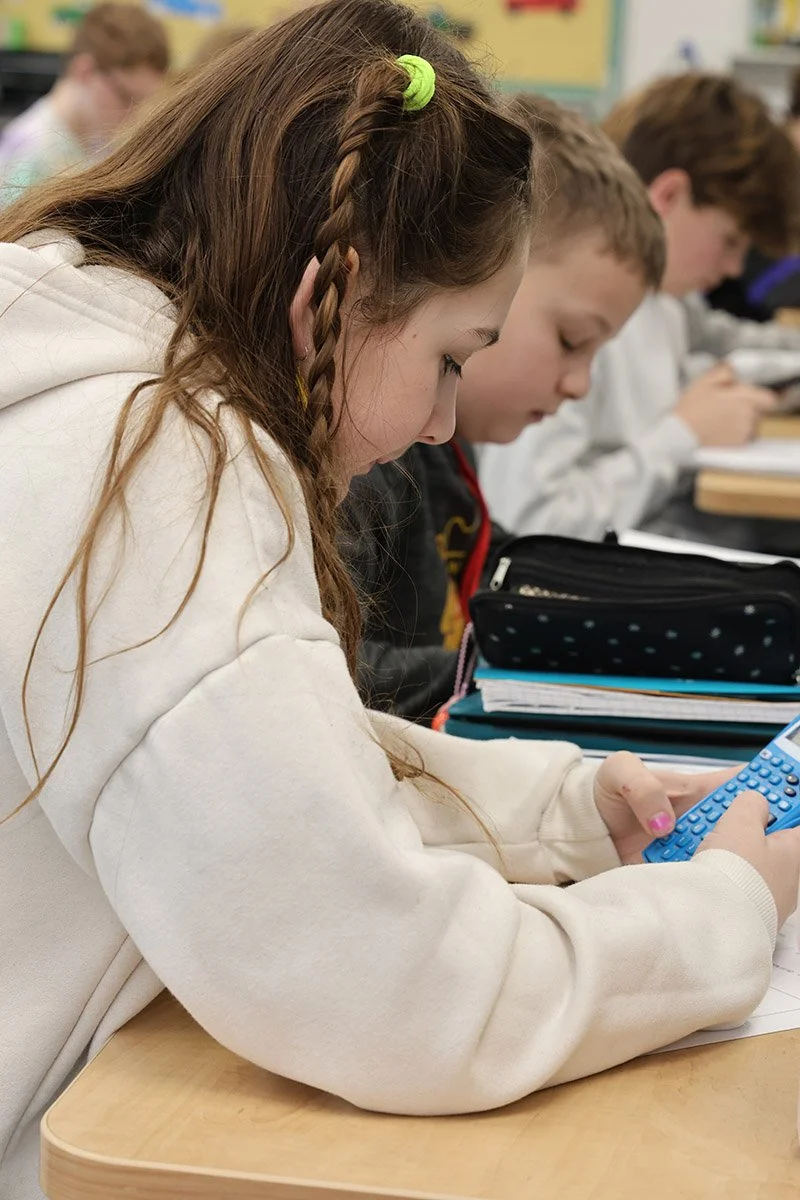 A girl with long brown hair, braid, wearing a white hoodie, is studying with a calculator in a classroom with other students.