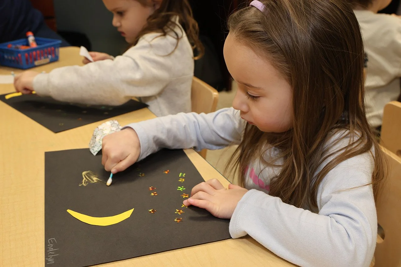 A young girl with long brown hair wearing a light gray long-sleeve shirt decorates a black paper with yellow sticker shapes and a sticker drawn face with a large smile, seated at a light wood table in a classroom.
