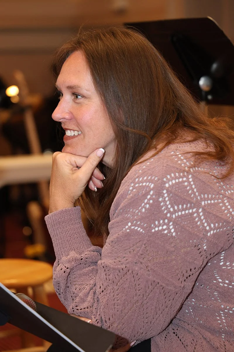 A woman with long brown hair, wearing a pink knitted sweater, smiling and resting her chin on her hand in a cozy indoor setting.