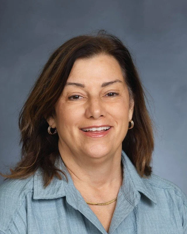 A  woman with shoulder-length brown hair, wearing a light blue shirt and gold jewelry, smiling at the camera against a gray background.