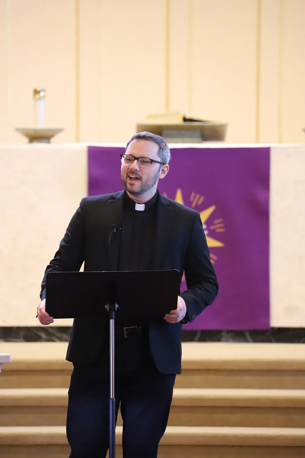 A man in a black clerical suit and glasses speaks at a lectern inside a church, with a purple and gold altar backdrop behind him.