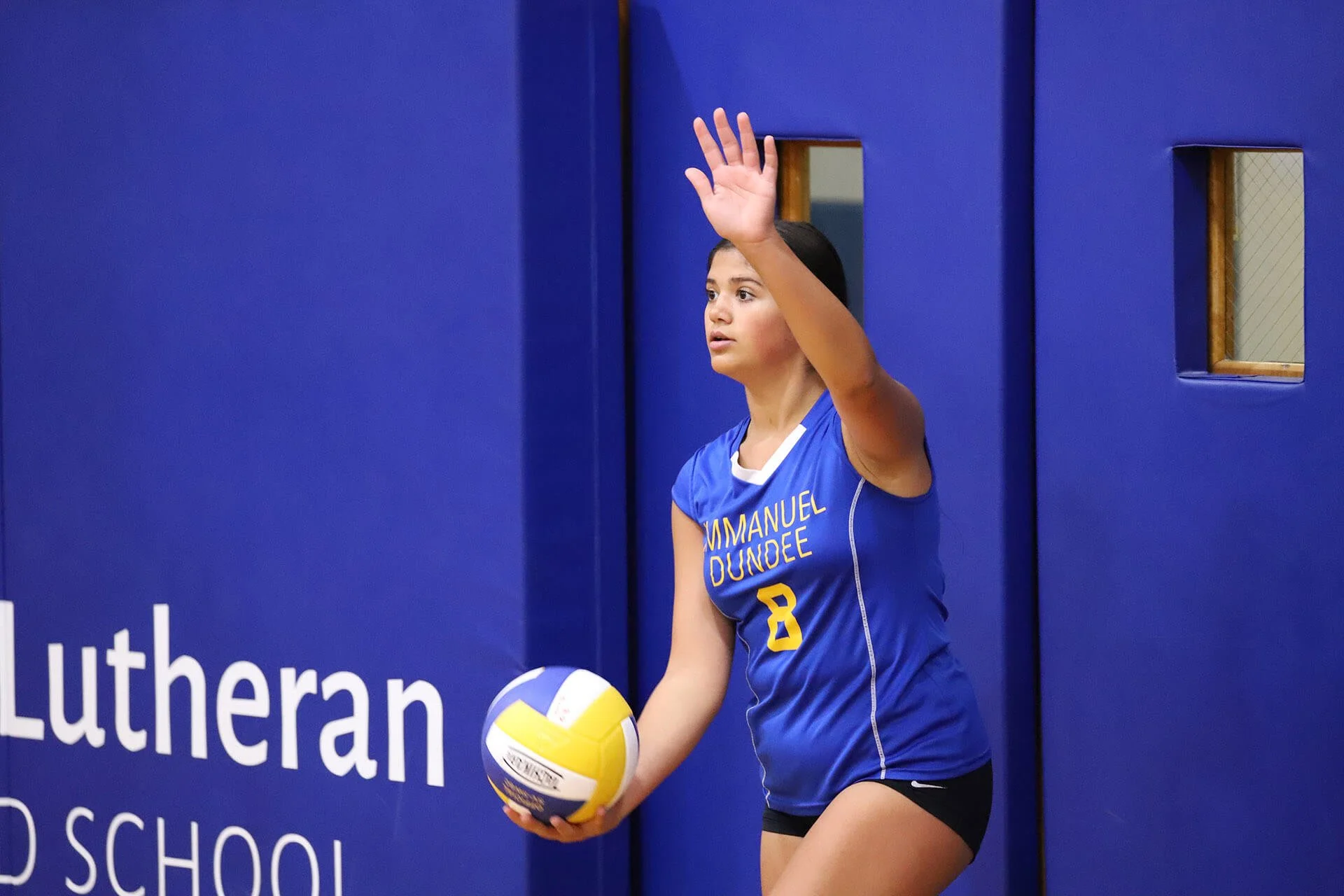 A female volleyball player in a blue and yellow uniform, holding a volleyball, with her hand raised near her head during a game.