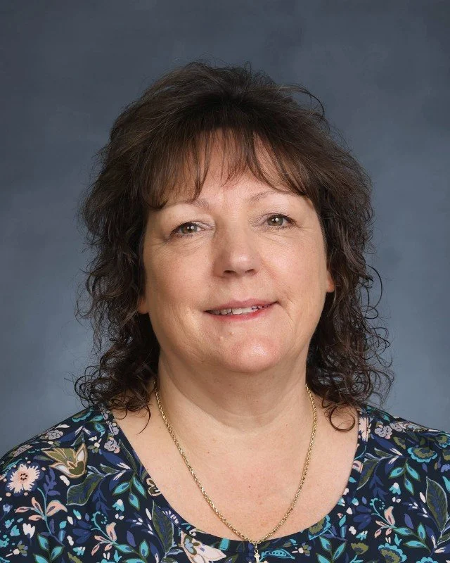 Portrait of a woman with shoulder-length curly brown hair, wearing a floral blouse and a gold necklace, against a blue-gray background.